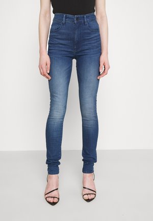 Jeans ajustados de tiro alto en azul, hechos de denim, con efecto desgastado, cinco bolsillos y cierre de botón, combinados con tacones negros de tiras.