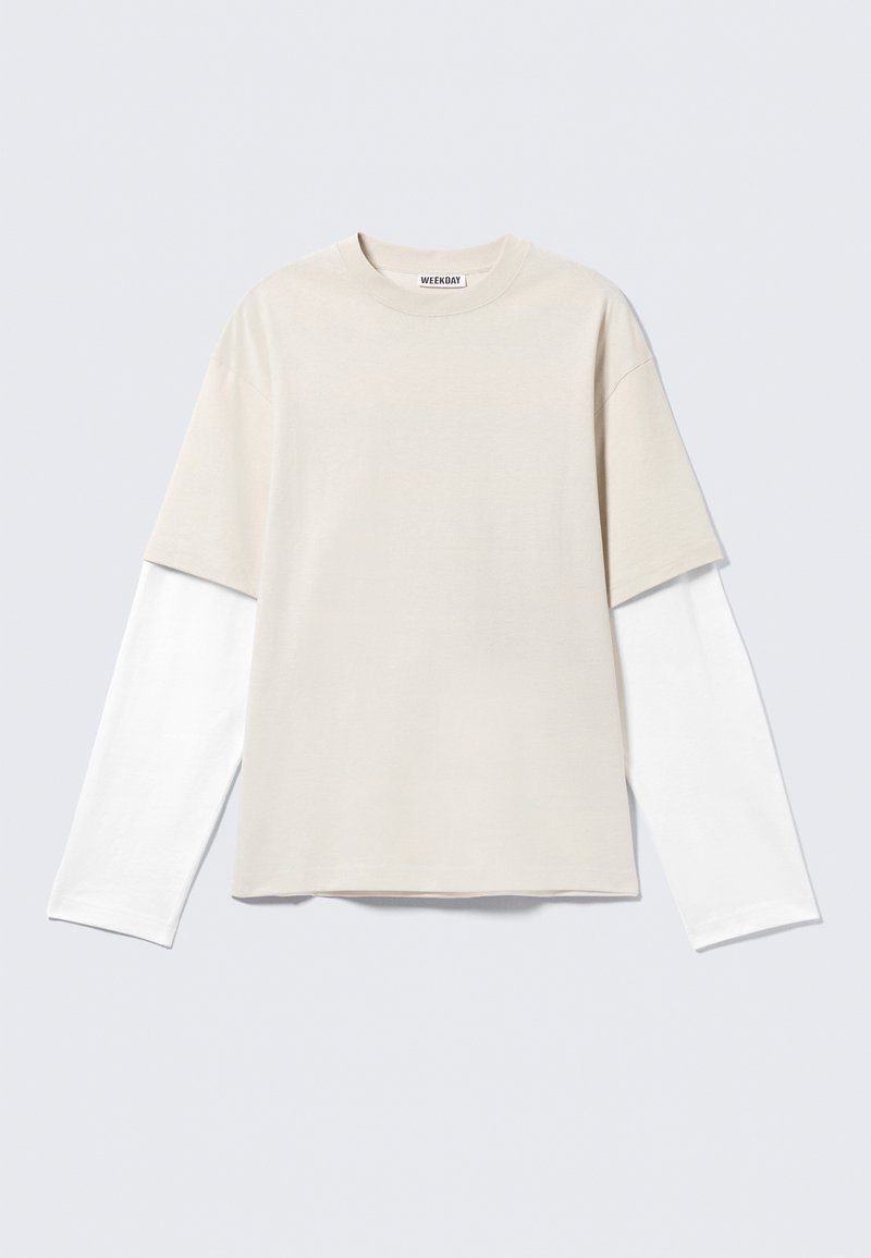 Μπεζ κοντομάνικο T-shirt φορεμένο πάνω από λευκή μακρυμάνικη μπλούζα με στρογγυλή λαιμόκοψη και ετικέτα "WEEKDAY" στο εσωτερικό του γιακά.