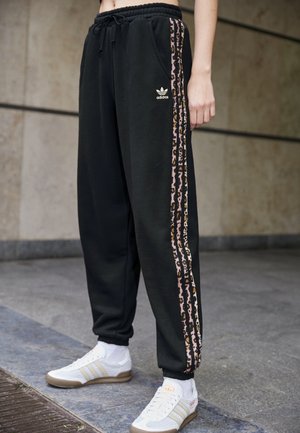 Osoba nosząca czarne dresy Adidas z lampasami w panterkę i białe sneakersy Adidas, stojąca na kafelkowanej podłodze.