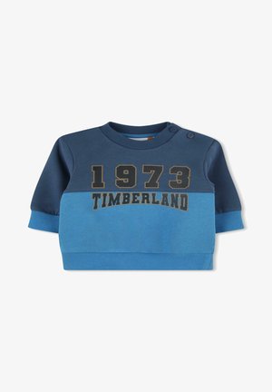 Sweatshirt bleu et bleu foncé à manches longues, avec des poignets côtelés, un col rond, et "1973 TIMBERLAND" imprimé en doré. Deux boutons-pression aux épaules.