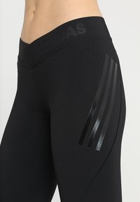 Leggings atletici neri con una texture liscia, dotati di una vita alta e tre strisce nere lucide sul lato sinistro.