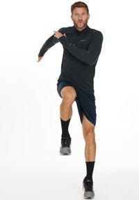 Aktive Herrenbekleidung bestehend aus einem langärmeligen, dunkelgrauen Shirt, navy Shorts, schwarzen Socken und grauen Sportschuhen. Laufpose.
