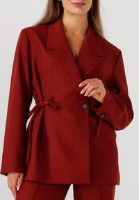 Bordeaux blazer met een getailleerde pasvorm, voorzien van een notched collar, dubbele knoopsluiting en gestrikte accenten aan de zijkanten. Soepele stoftextuur.