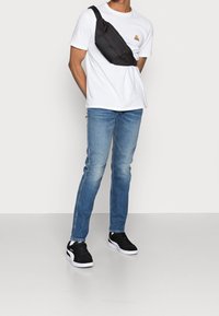 Svart axelväska med texturerad framsida, bärs över en vit t-shirt, matchas med ljusblå jeans och svarta sneakers. Enkel och funktionell design.