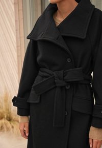 Palton negru din lână cu guler larg, nasturi mari și curea legată. Include buzunare aplicate și mansete pliate cu un tricot texturat dedesubt.