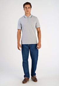 Grå polo t-shirt med mörka detaljer på kragen och ärmarna, kombinerad med blå jeans och bruna båtskor, presenteras i en stående pose.