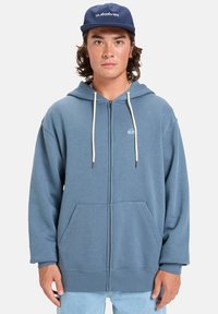 Sweat à capuche zippé bleu en tissu doux, avec une capuche, des poches kangourou et des cordons blancs. Inclut un petit logo sur la poitrine.