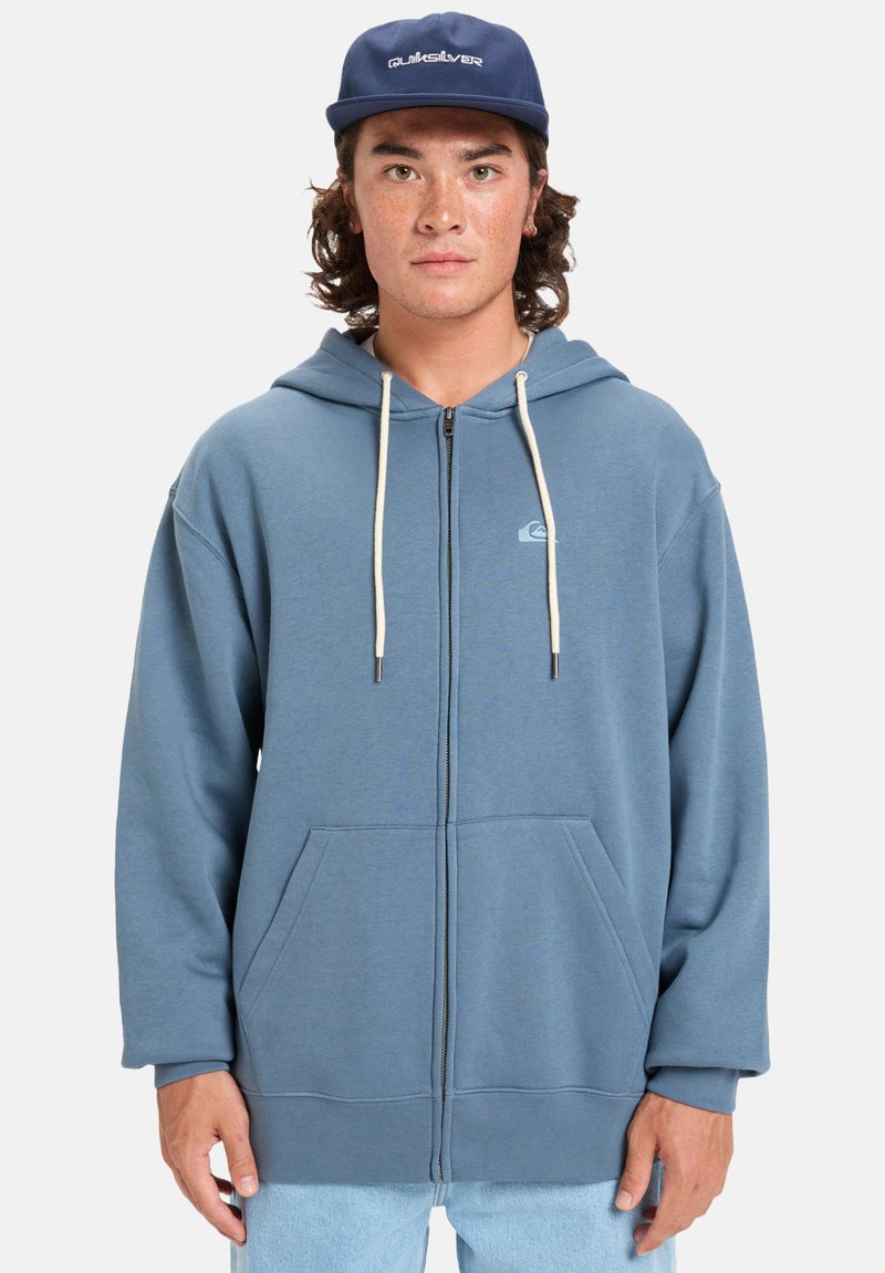 Sweat à capuche zippé bleu en tissu doux, avec une capuche, des poches kangourou et des cordons blancs. Inclut un petit logo sur la poitrine.