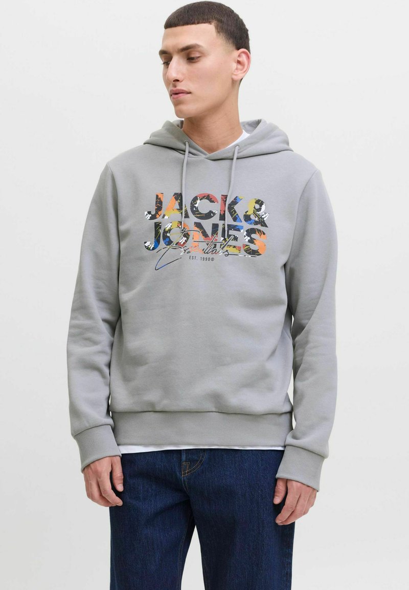 Felpa grigia con cappuccio e coulisse, caratterizzata da una stampa grafica multicolore "JACK & JONES" e polsini e orlo a costine, indossata sopra dei jeans.