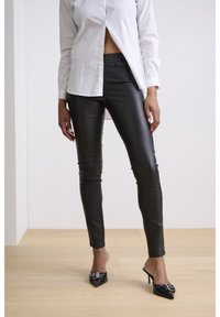 Czarne legginsy z ekoskóry z elastycznym pasem, o dopasowanym kroju i błyszczącej fakturze; zestawione z białą koszulą na guziki i czarnymi mulesami na obcasie.