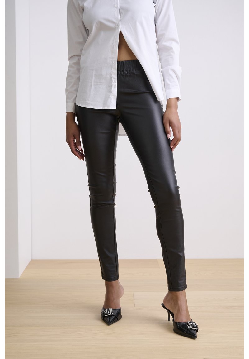 Czarne legginsy z ekoskóry z elastycznym pasem, o dopasowanym kroju i błyszczącej fakturze; zestawione z białą koszulą na guziki i czarnymi mulesami na obcasie.