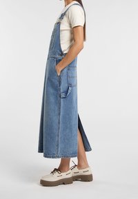 Denim overall šaty v světle modré barvě, s bočními kapsami, nastavitelnými ramínky a vysokým rozparkem. Nosené s obuví v bílé barvě.