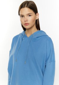 Felpa azzurra chiara, con cappuccio regolabile, vestibilità ampia e texture del tessuto morbida. I dettagli includono accessori in metallo argentato e un design minimale.