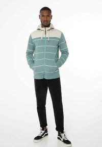 Lichtblauwe ritshoodie met crèmekleurige horizontale strepen, voorzakken, verstelbare capuchon, gecombineerd met zwarte broek en zwarte sneakers.