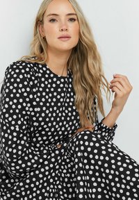 Vestido negro con grandes lunares blancos, mangas largas, escote redondo y tejido fluido, que presenta una textura suave y un drapeado delicado.