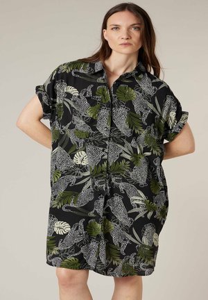 Femme portant une robe noire boutonnée ornée de feuilles vertes et d'un imprimé léopard blanc à pois, debout les mains derrière le dos.