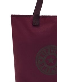 Maroon tote bag with a smooth texture, featuring a logo in a darker shade and a sturdy fabric handle. Simple design with no embellishments.