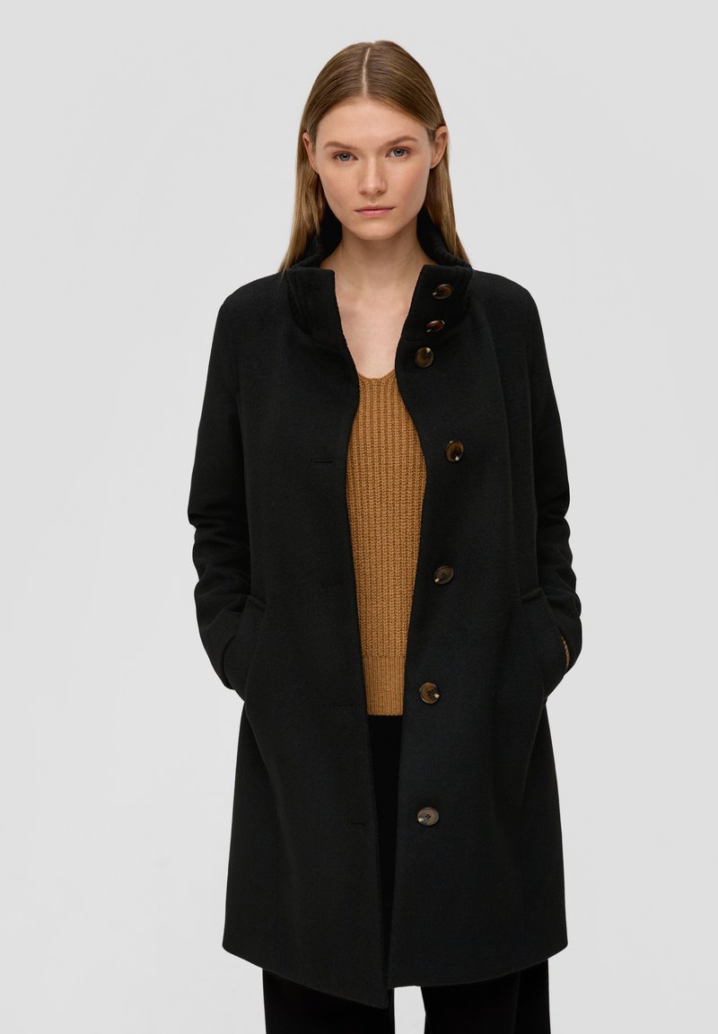 Manteau noir en laine avec un col haut et une fermeture à boutons, doté de deux poches latérales et d'un pull en tricot brun texturé en dessous.