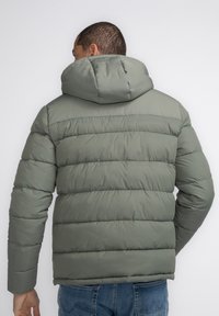Chaqueta acolchada verde con capucha, que presenta una textura acolchada, puños elásticos y una forma cuadrada. Material suave y diseño simple.