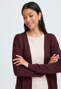 Cardigan bordeaux con design a apertura frontale, maniche a coste e tasche, indossato sopra un maglione leggero a coste. Texture liscia, vestibilità accogliente.