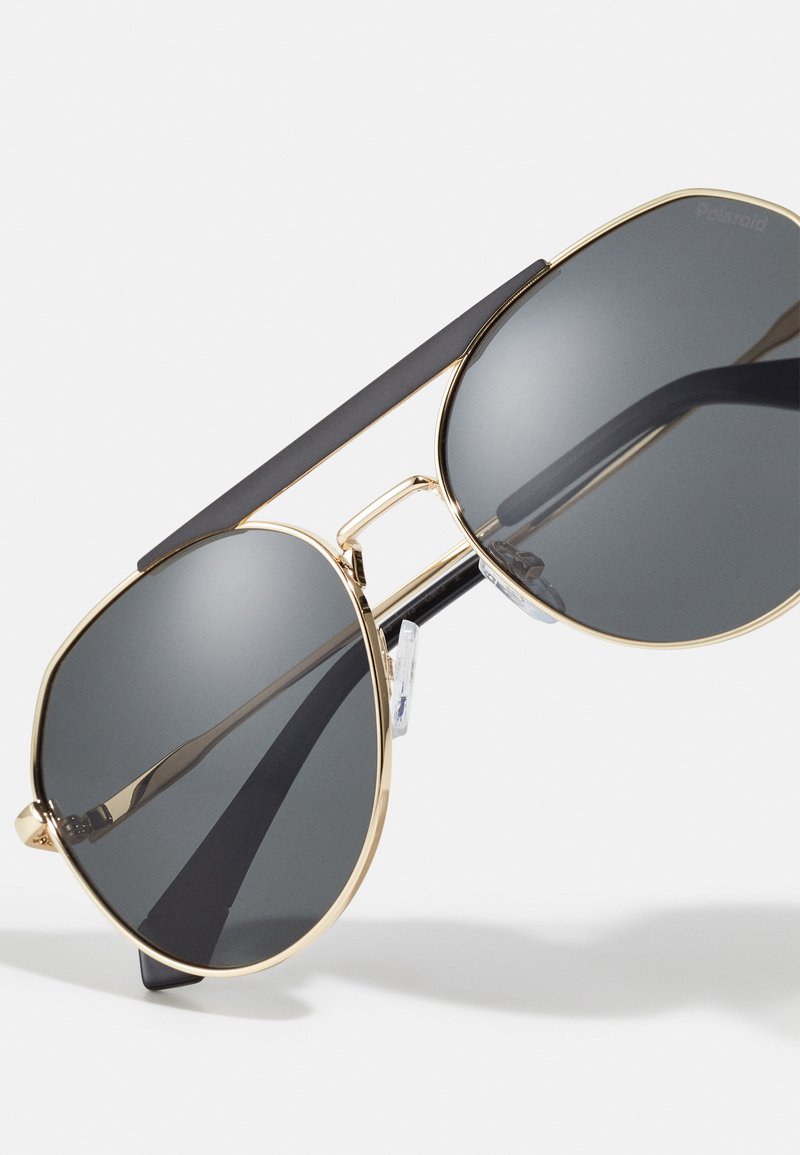 Gafas de sol aviador de metal dorado con lentes gris oscuro, acentos negros en la parte superior y almohadillas transparentes en la nariz. Presenta un diseño elegante y minimalista.