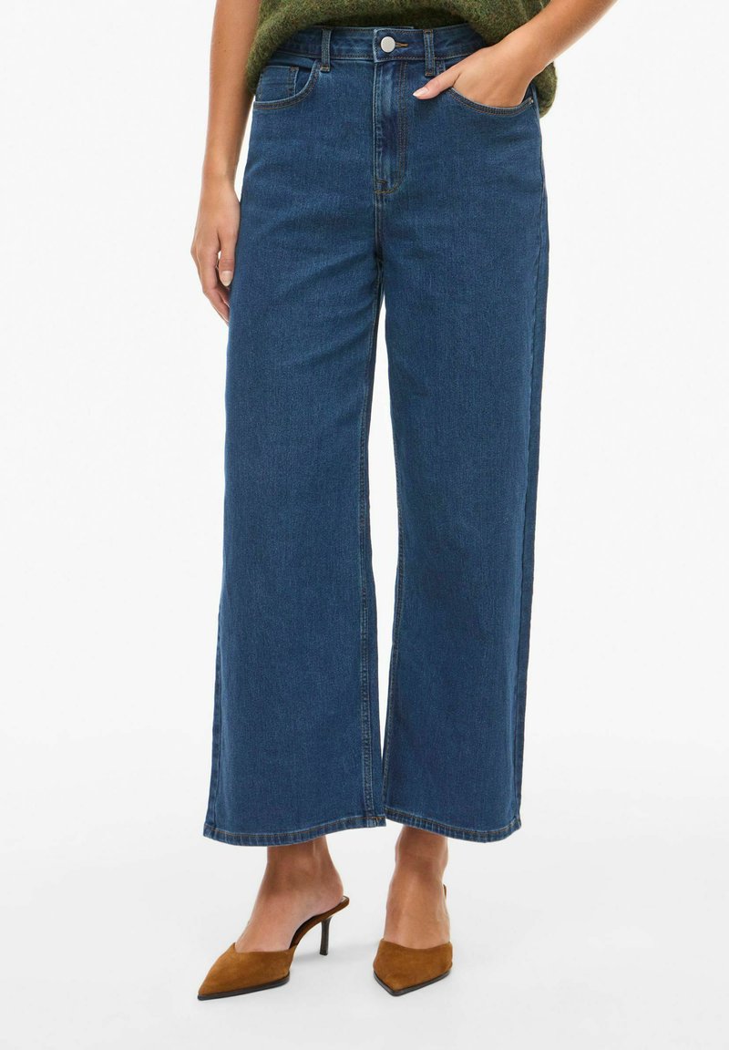 Jeans de mezclilla azul de pierna ancha con cintura alta, bolsillos frontales y cierre de botón. La tela tiene una textura suave y un acabado clásico.