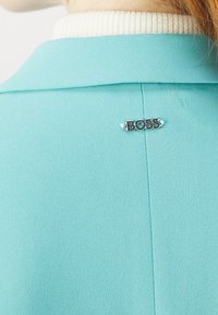 Tyrkysový blazer s jemnou texturou, který má bílý ribovaný límec. Na zadní straně je umístěna kovová brož s logem "BOSS".