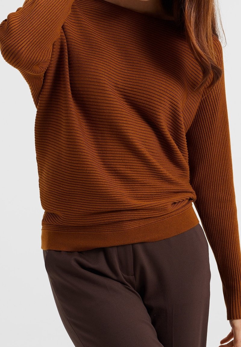Femme portant un pull moulant côtelé à manches longues de couleur rouille et un pantalon marron foncé sur un fond uni.