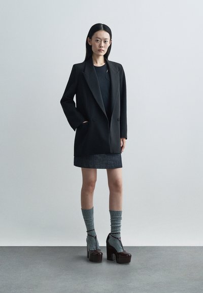 Czarna podwójnie zapinana marynarka z dużymi klapami, w połączeniu z ciemną spódnicą, szarymi prążkowanymi skarpetkami i butami na masywnym obcasie. Czysta, nowoczesna sylwetka.