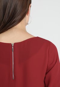 Blusa rossa con scollo rotondo e maniche corte. Dettagli con zip sul retro in finitura metallica argento. Tessuto dalla trama liscia.