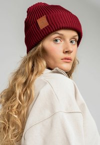 Bordowa, dziana czapka z pionowym karbowaniem, z brązową skórzaną łatą. Miękki, fakturowany materiał o luźnym kroju.