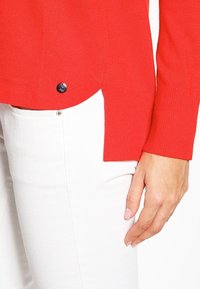 Höger hand bredvid fickan på de vita byxorna, iförd en röd långärmad tröja med en liten rund metalldetalj nära fållen.