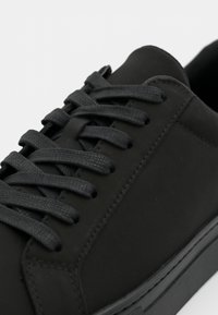 Μαύρο αθλητικό παπούτσι από λείο δέρμα με επίπεδες μαύρες κορδόνες, διακριτική ραφή και σόλα χαμηλού προφίλ.