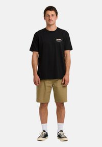 Zwarte katoenen t-shirt met een klein logo op de borst, gecombineerd met kaki shorts en zwarte sneakers met witte zolen en veters.
