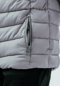 Grå vadderad jacka med quiltad struktur, horisontella paneler och en sidoförslutning med texturerad dragflik. Materialet verkar mjukt och lätt.