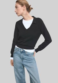 Maglione nero a maglia con scollo a V e colletto, indossato sopra una camicia bianca a maniche lunghe, abbinato a jeans a gamba larga di un blu chiaro.