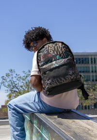 Mochila negra y gris con diseños gráficos, que presenta un ciclista de BMX y patrones abstractos. Fabricada con tela duradera, cuenta con correas ajustables.