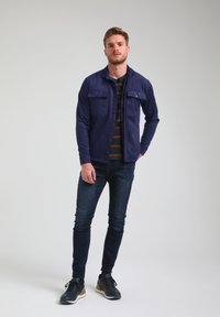 Giacca navy in suede con due tasche sul petto, chiusura a bottone e finitura testurizzata, indossata sopra una camicia a righe e jeans scuri con sneakers.