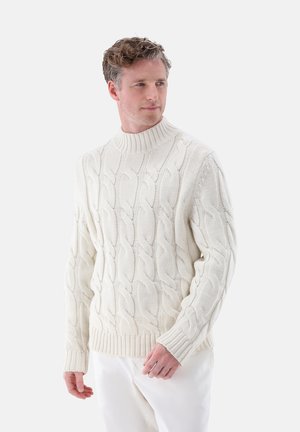 Fettes cremefarbenes Strickpullover mit hochgezogenem Rippkragen und Rippbündchen; getragen über weißen Hosen mit strukturierter Optik.