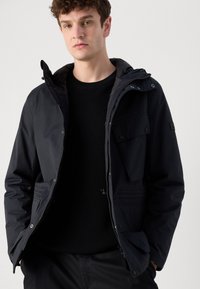 Veste noire imperméable avec capuche, fermeture éclair et poches avant doubles. Présente une texture lisse et un design ajusté.