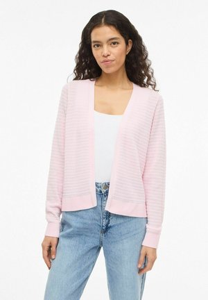 Donna con capelli ricci che indossa un cardigan aperto di colore rosa chiaro con texture sopra una maglietta bianca e jeans blu, in piedi davanti a uno sfondo bianco.
