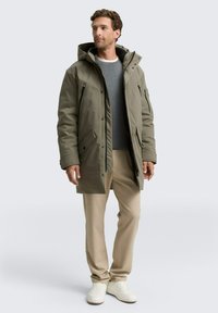 Olivno zelena parka s kapuco, zadrga spredaj, veliki stranski žepi in gladka tekstura. Nošena čez sivo jopico in bež hlače.