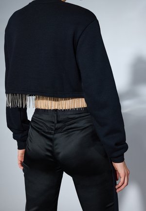 Personne portant un sweat court noir avec des détails en franges argentées et un pantalon en satin noir taille haute, vu de dos.