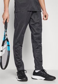 Donkergrijze sportbroek met geometrische patronen, met een slim fit en elastische tailleband. Gecombineerd met zwarte sneakers en een tennisracket.