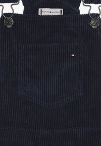 Tmavě modré manšestrové kalhoty na kšandy s horizontálním žebrováním, nastavitelnými kovovými sponami a malou kapsou s červeno-bílo-modrým logem.