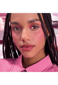 Femme aux cheveux foncés tressés portant un fard à paupières rose, du blush et du rouge à lèvres, vêtue d'une chemise rose à col, sur un fond de nuages roses.