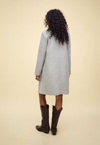 Gray wool coat with long sleeves, mid-length design, and a straight silhouette, paired with brown cowboy boots. Background is beige.