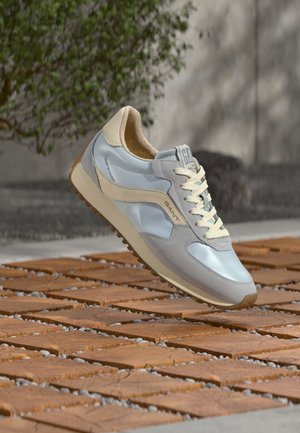 Pantof sport GANT albastru deschis și bej, cu șireturi albe, plutind deasupra unei pavaje din blocuri de lemn în aer liber, lângă verdeață.