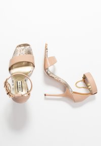 Nude högklackade sandaler med en glänsande lackfinish, texturerade metalliska detaljer och ett smalt ankelsnöre med kedjedetalj.