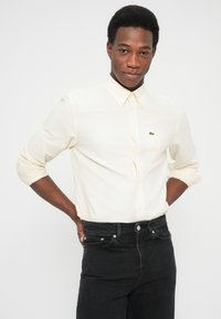 Hombre con los brazos cruzados, vistiendo una camisa crema abotonada con las mangas remangadas y jeans negros, sobre un fondo blanco.